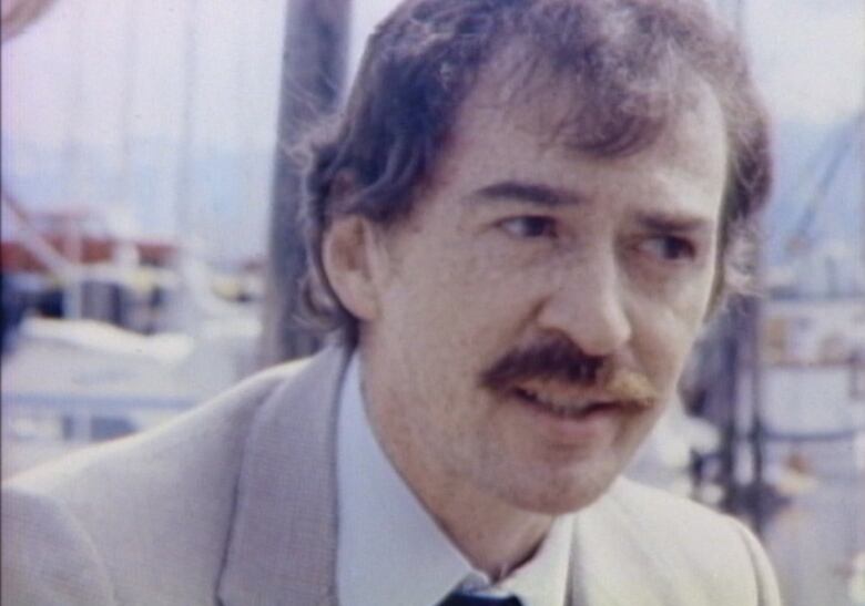 A close up of a man in a coat and tie sitting at a marina.