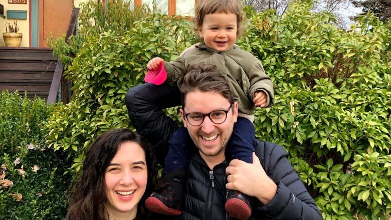 A man with a toddler on his shoulders, and woman next to him, all smiling.