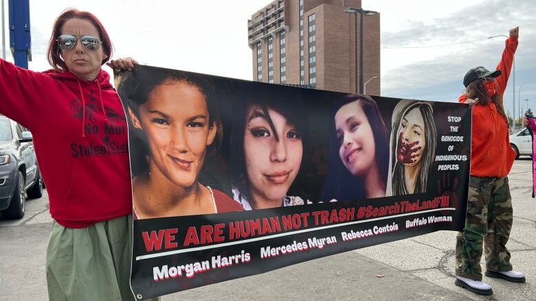 Two women wearing red sweatshirts hold a poster that says 'we are human not trash hashtag search the landfill' with photos of four murdered Indigenous women. 