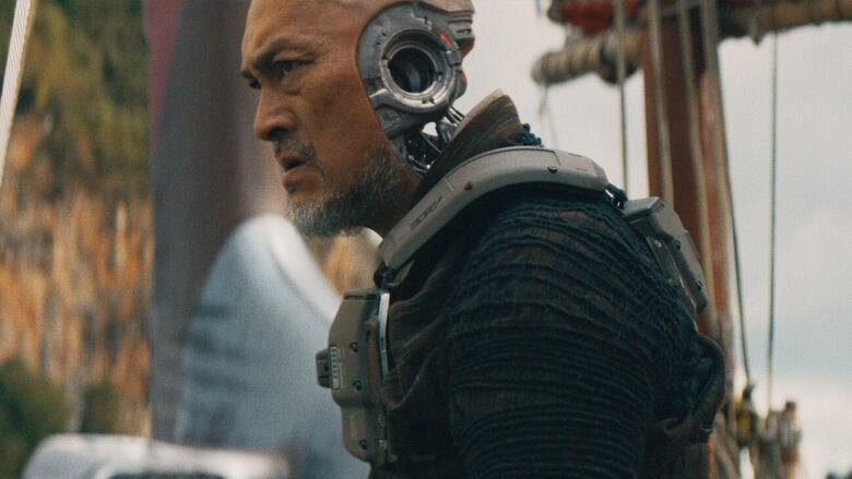 A man wearing a futuristic, mechanical vest scowls. Where his ear should be is a mechanical structure mimicking a skeleton, with a hole going through the centre. 