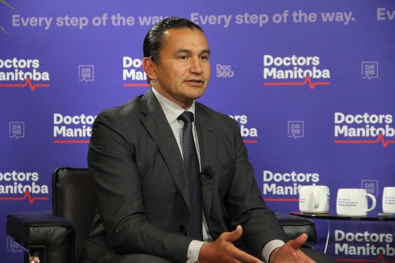 A man in a suit and blazer speaks while seated in a chair.