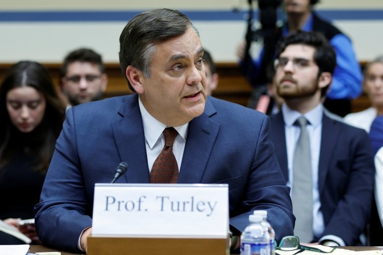 A man in a suit and tie speaks while seated at a table.