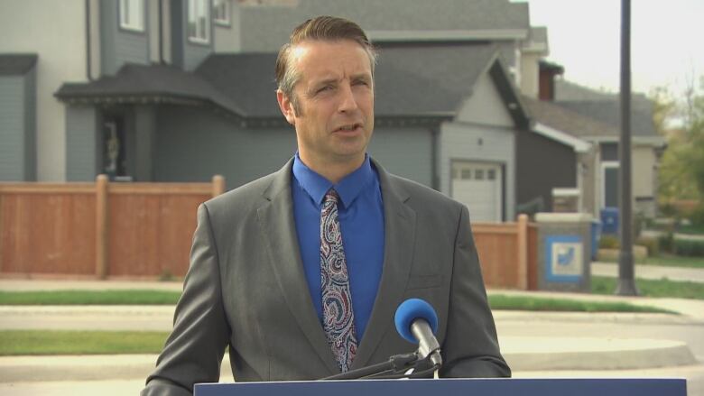 A man in a suit and tie speaks at a podium in front of houses in a residential area.