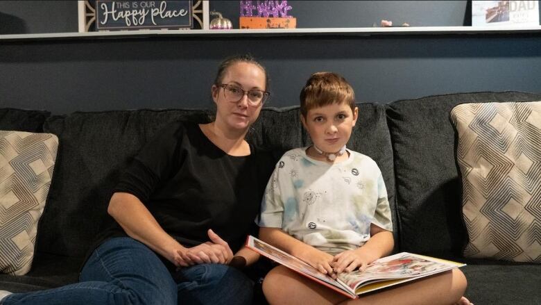 A mother and son sit on a couch. The son has a book on his lap and a small medical device around his neck.