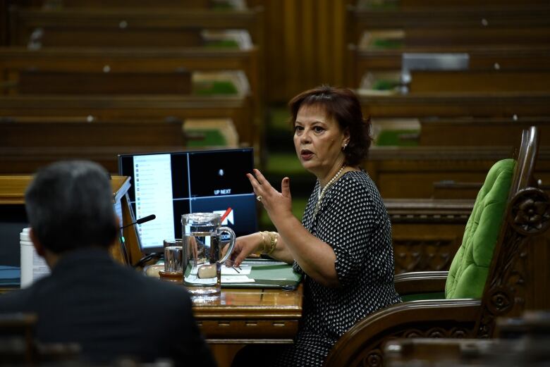 A woman sits at a desk in the House of Commons, her left arm raised.