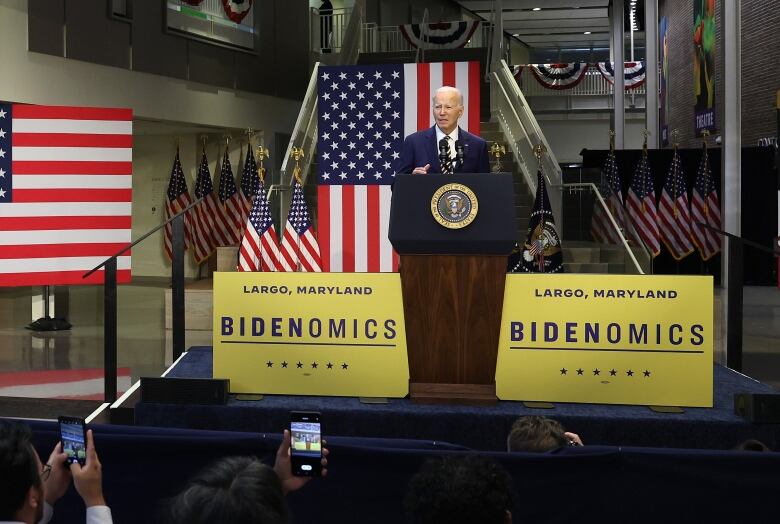 Joe Biden stands at a podium set up on a small stage in an auditorium, flanked by American flags and yellow posters with the words 'Bidenomics' and 'Largo, Maryland.' 