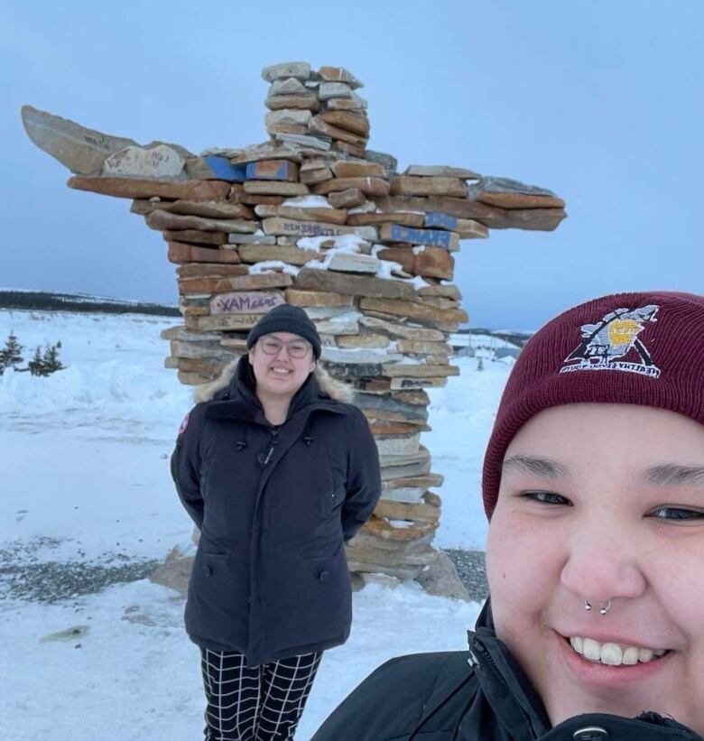 an Indigenous couple smiling to pose in front of am Inuksuk, a figure made of piled stones. 