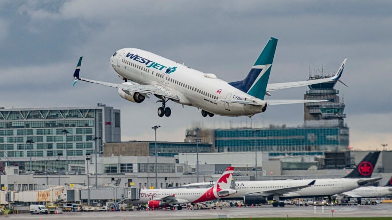 Planes on tarmac in foreground, Westjet plane taking off. Airport building in background.