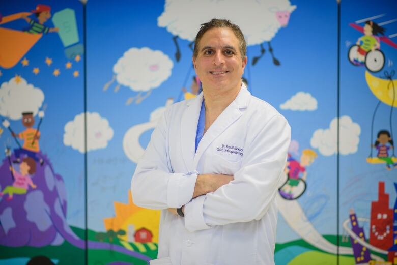 Man in white doctor's coat stands in front of a mural of children playing. 