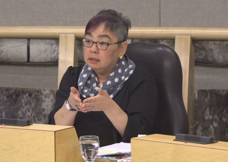 A woman with short hair and a scarf sits in the legislative assembly and presents. 