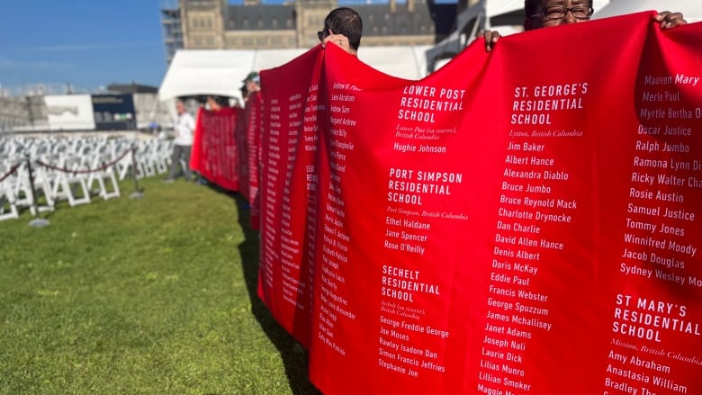 A line of people standing on a lawn hold a long red banner, bearing the names of people and residential schools. 