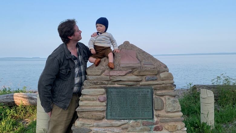 A little boy sits on a stone cairn replica bearing a plaque. His father stands nearby.