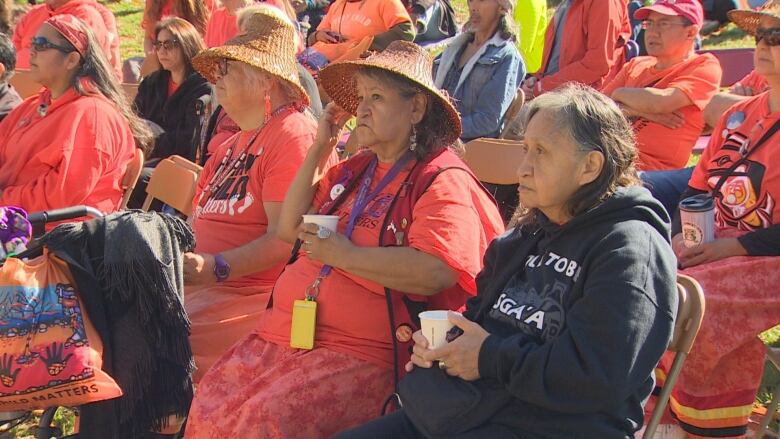 A group or Indigenous elders wearing orange shirts sit in chairs outdoors.