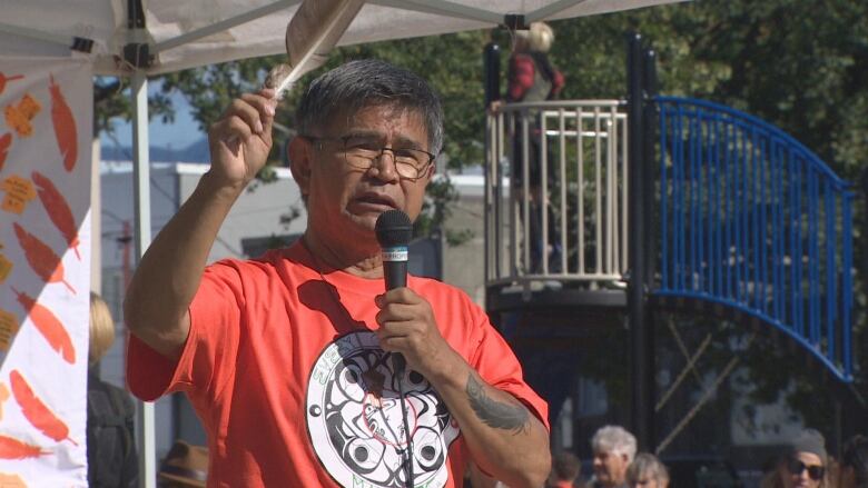 An older man in an orange shirt holds a feather and speaks in a mic.