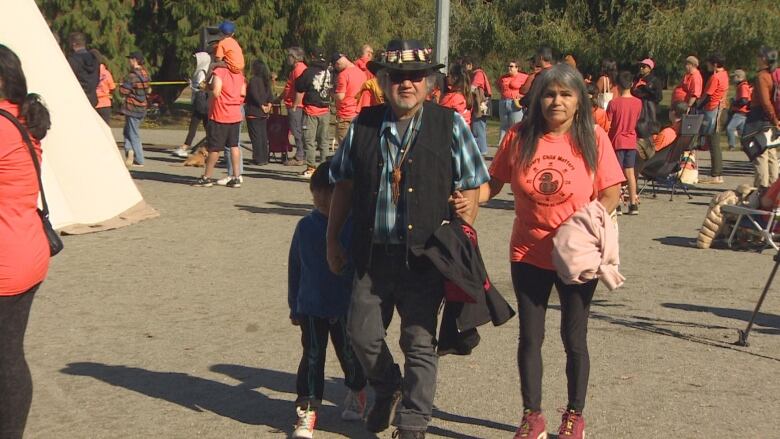 An older couple in orange shirts walk with a crowd and tipi behind them.