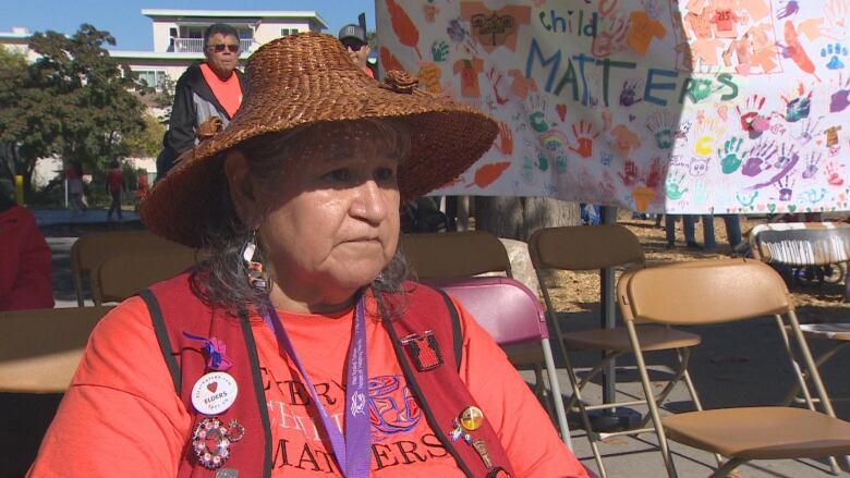 An older woman in a cedar straw hat speaks to the camera.