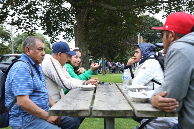 Four men sitting on a table, listening to a woman speaking.