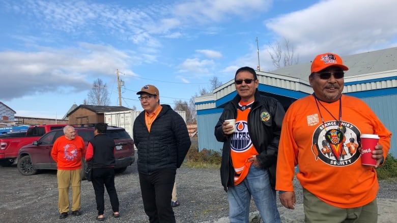 Five people on a sunny day standing behind a fire in front of a building. The two men on the right smile. Both are wearing orange.