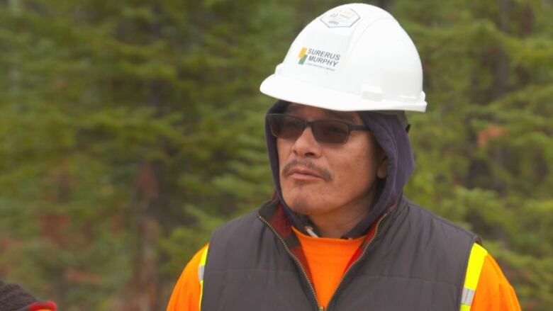 Frank Roan, a representative from Mountain Cree, wears a white hard hat, brown hoodie and reflective vest. He stands on a job sit in Jasper, Alberta.