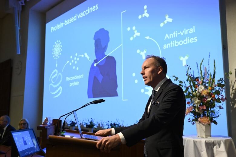 A man is shown speaking at a podium from a side view on a stage, with a large projected screen behind him.