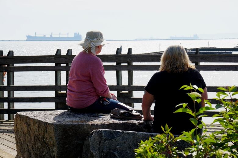 Two women gaze out at the water, which has big ships sailing on them. 