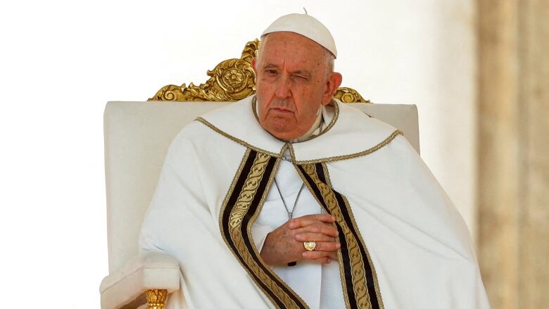 An older man in a white cape lined with black and gold embroidery sits on a throne with his hands crossed.