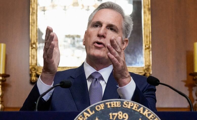 Grey haired man gestures with two arms as he speaks in microphones