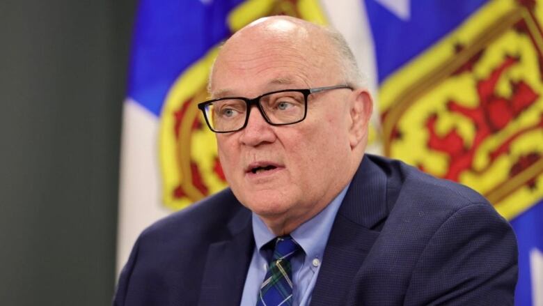 A man with glasses and wearing a blue jacket, shirt and tie, speaks in front of Nova Scotia's provincial flags.