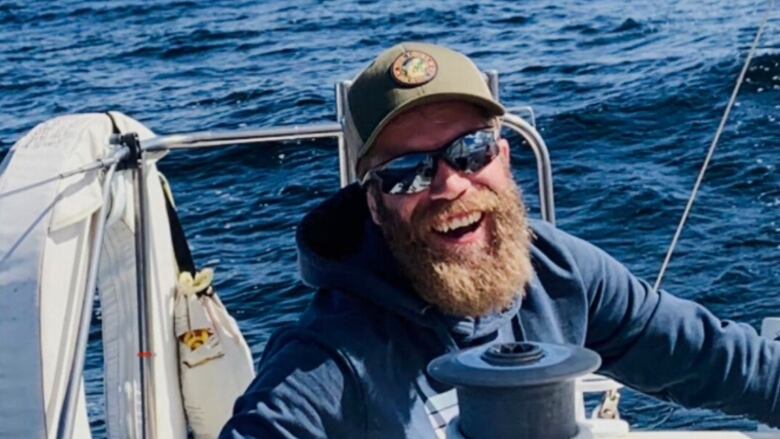 A man with a brown beard and a hat and a blue jacket is on a boat, surrounded by water.