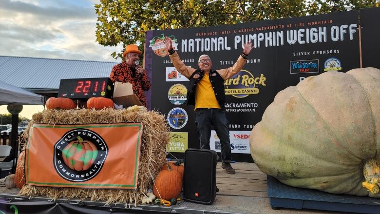 A man stands cheering on a stage in front of a giant pumpkin that reaches his chin in height. 