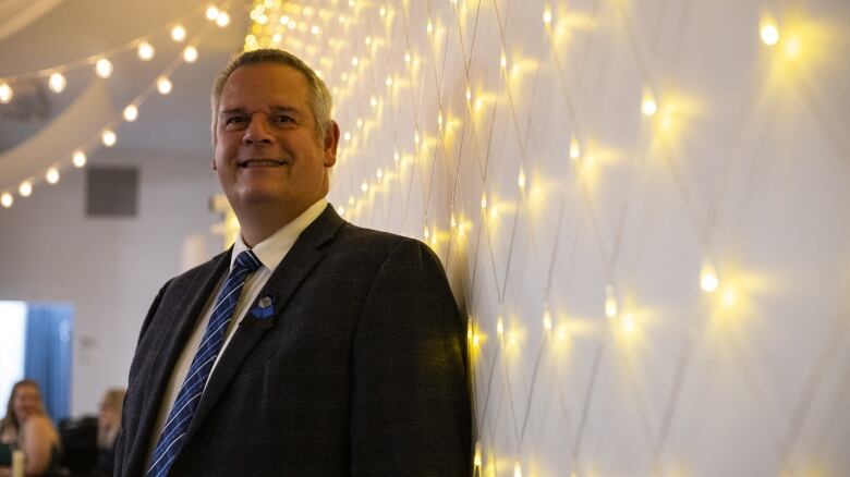 A man stands by a lit up wall wearing a Progressive conservative pin.