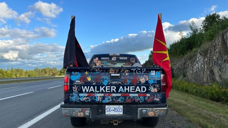 A truck covered in handprints with the words WALKER AHEAD printed across the back sits parked on the side of a highway. Two flags stick up out of the truck bed. 