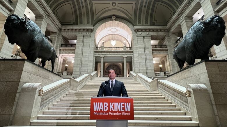 A man stands behind a podium that says Wab Kinew. Behind him is a grand staircase with large bison statues at either side.