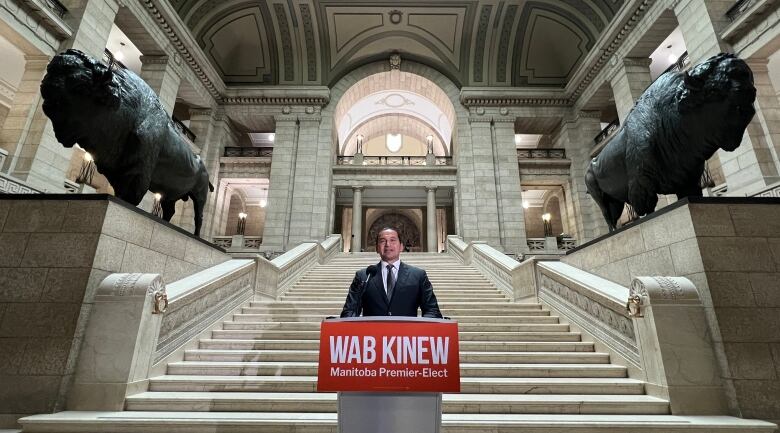 A man stands behind a podium that says Wab Kinew. Behind him is a grand staircase with large bison statues at either side.