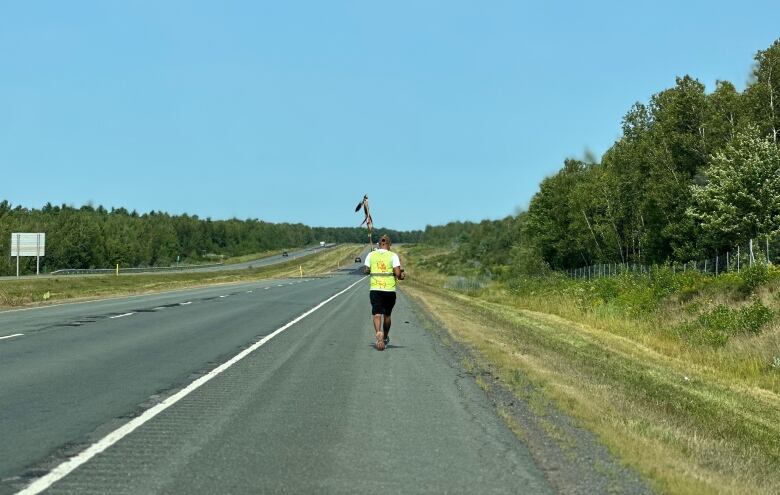 A man holding a staff and wearing a yellow safety vest walks down a highway.