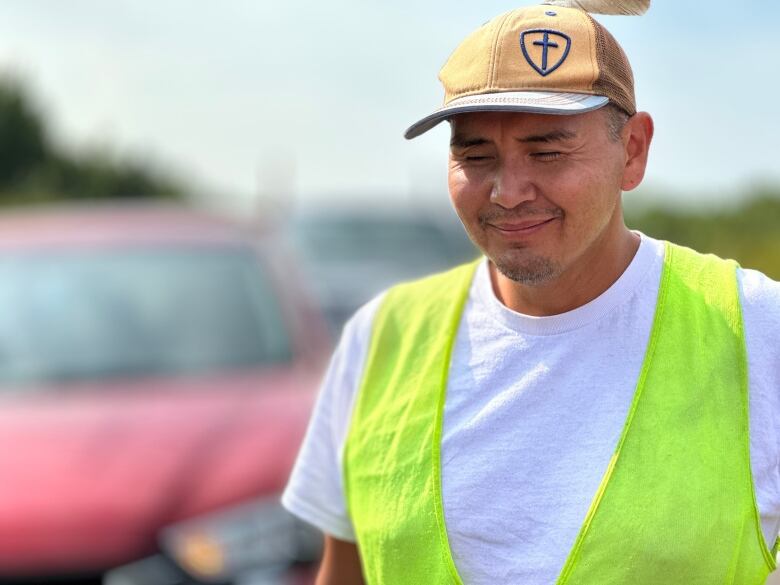 A smiling man wearing a white t-shirt and yellow safety vest stands outside in front of a red car.