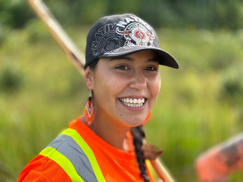 A smiling woman wearing a baseball cap and orange earrings and a safety vest stands outside.