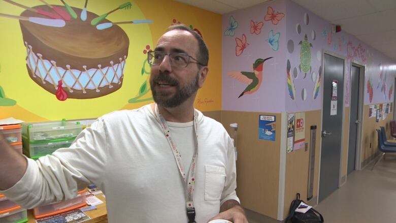 A man in a long-sleeved white T-shirt stands in front of a brightly coloured mural with Indigenous motifs.