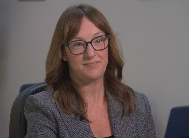 A woman with glasses and shoulder-length hair wearing a blazer sits on a chair in a boardroom.