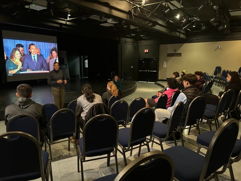 A man stands in front of a group of people sitting in chairs. A projector behind him shows an image of a man in a suit speaking at a podium.