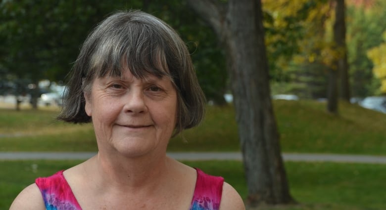 A woman in a colourful tanktop stands in front of some trees