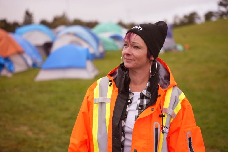 A woman in a black toque and an orange and yellow reflective jacket. She's standing in a green field with tents behind her.