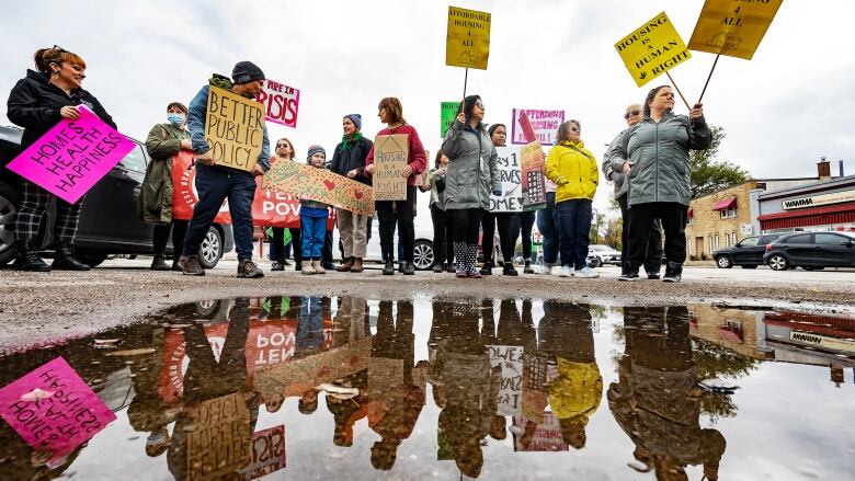 A dozen people hold signs demanding more affordable housing. The crowd is reflected in a puddle on the ground.