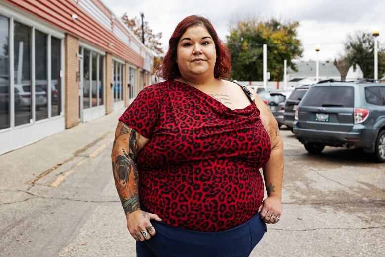 A woman stands in a parking lot. She wears a red leopard print shirt and blue pants. She looks concerned.