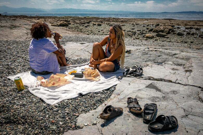 Two women sit across from each other on a blanket spread out on a beach. 