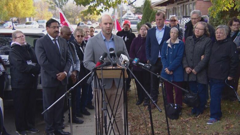 A man wearing a grey suit is standing at a podium with microphones on it as he delivers a press conference, he is surrounded by people standing behind him. 