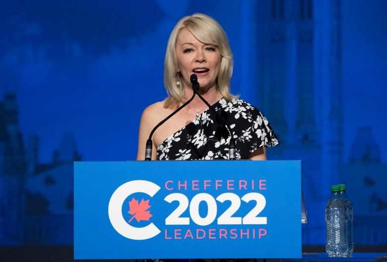 A woman wearing a patterned dress speaks at a podium with a sign bearing the Conservative Party's logo and reading 