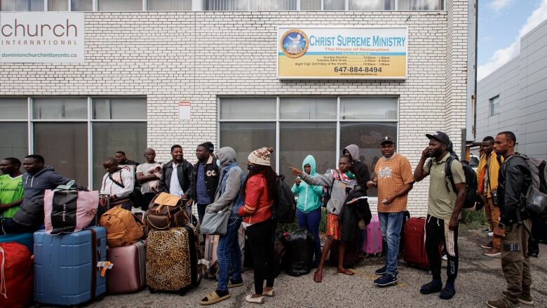 A line of Black asylum-seekers in Toronto with their luggage, waiting to board a chartered bus.