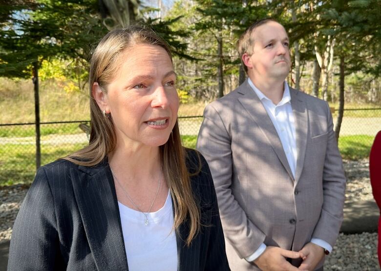 A blonde woman with a grey jacket and white shirt speaks as a man in a grey suit looks on.