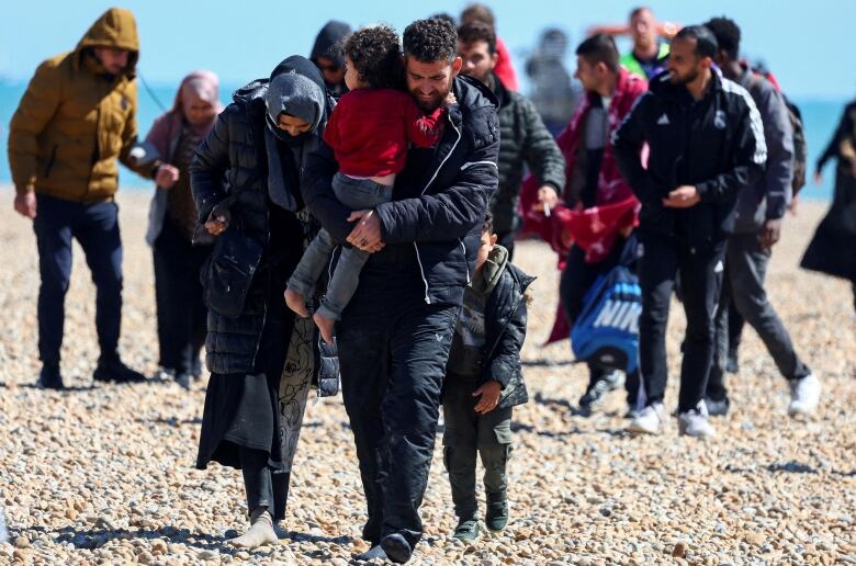 A man carries a child as a large group of people walk on a stoney shore.